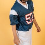 Future Skills - Full length Asian boy in American football player uniform with number and helmet standing in studio on yellow background and looking away