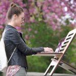 Workplace Flexibility - Woman in Leather Jacket Standing on a Park while Working on Her Laptop