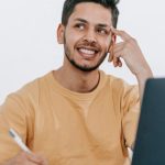 Freelancing Future - Smiling young bearded Hispanic male entrepreneur thinking over new ideas for startup project and looking away dreamily while working at table with laptop and taking notes in notebook