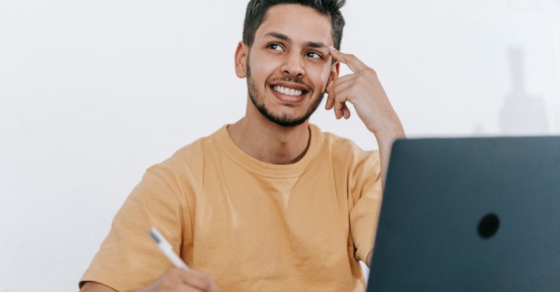 Freelancing Future - Smiling young bearded Hispanic male entrepreneur thinking over new ideas for startup project and looking away dreamily while working at table with laptop and taking notes in notebook