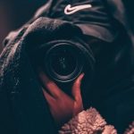 Hidden Jobs - Unrecognizable person wearing hooded clothes and hiding face behind black scarf taking photo on professional camera on blurred background