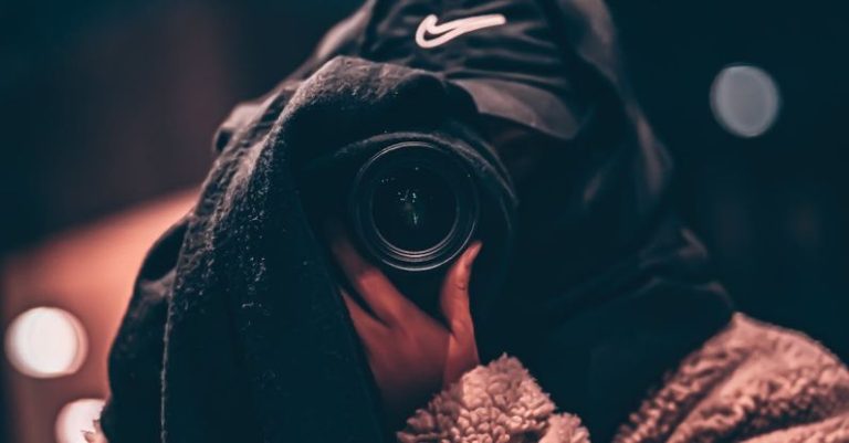 Hidden Jobs - Unrecognizable person wearing hooded clothes and hiding face behind black scarf taking photo on professional camera on blurred background